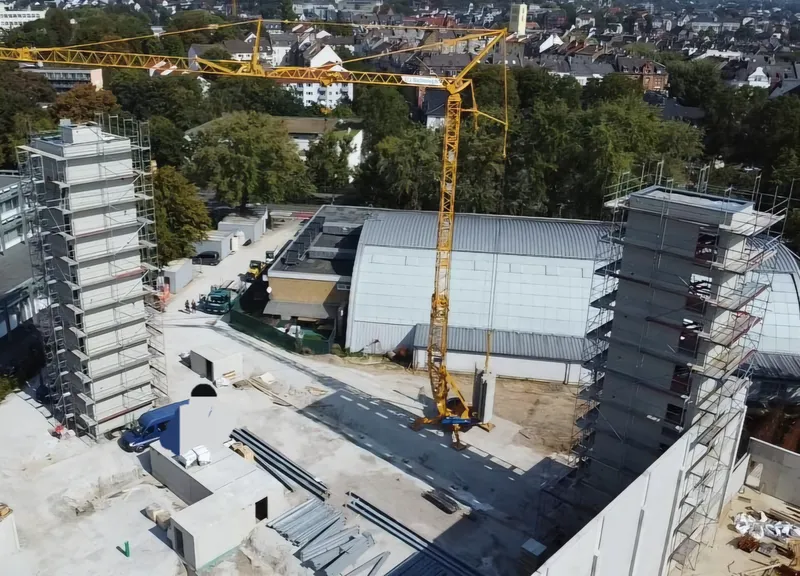 Luftaufnahme einer Baustelle: zwei eingerüstete Gebäudekerne, Baukran, Halle mit Rundbogendach