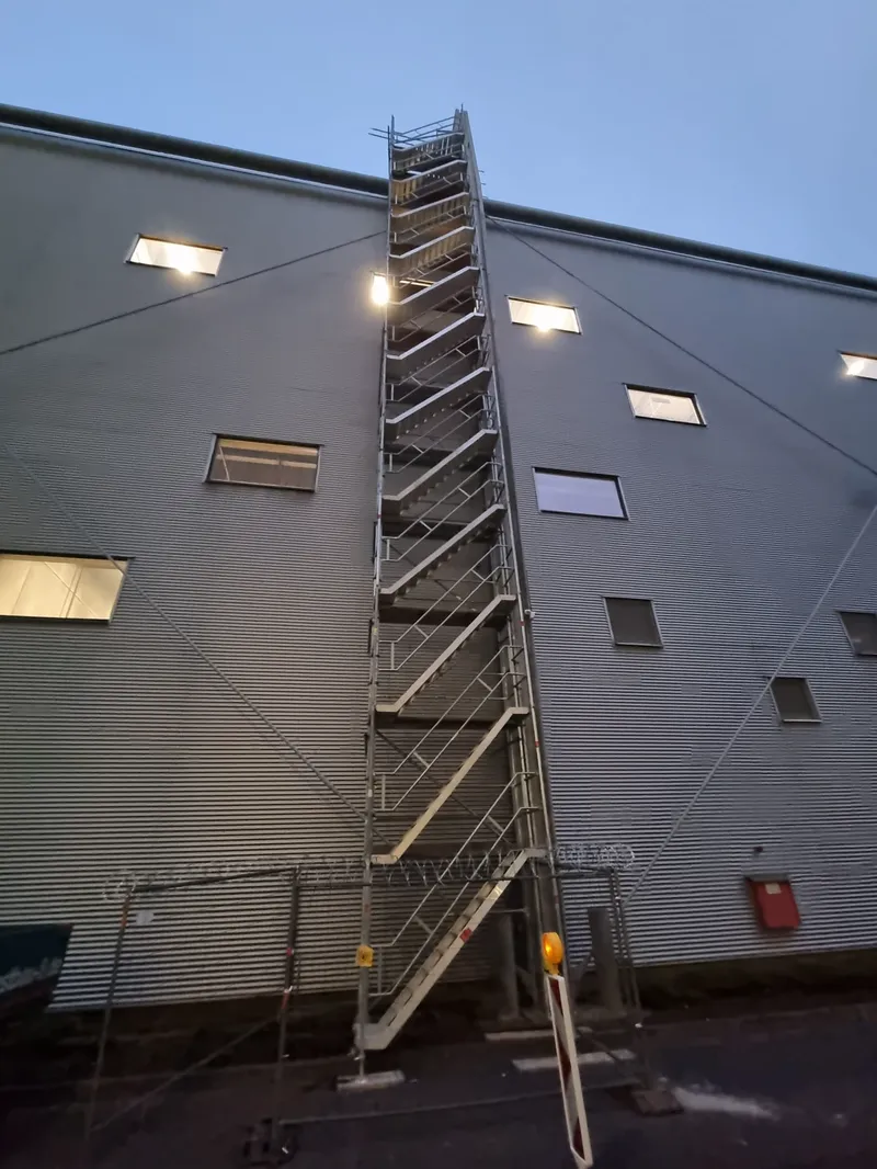 Treppenturm an der Fassade einer Industriehalle in der Dämmerung, Bauzaun davor