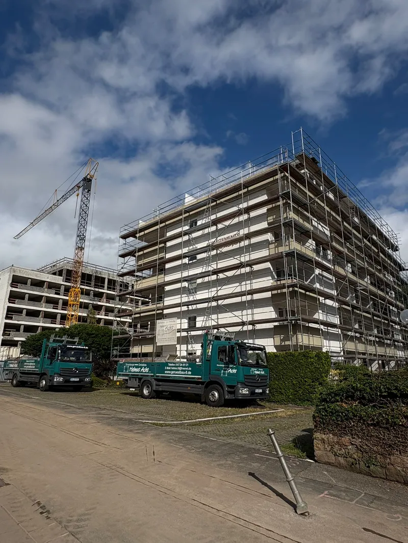 Dasselbe Appartementhaus mit Baukran und zwei Firmen-LKW, daneben ein Rohbau