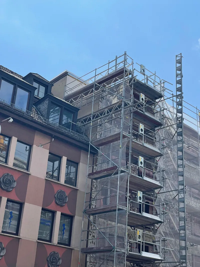 Innenstadtfassade mit Rosetten, eingerüstet mit Bauaufzug und Netz, daneben ein Schieferdach mit Gauben