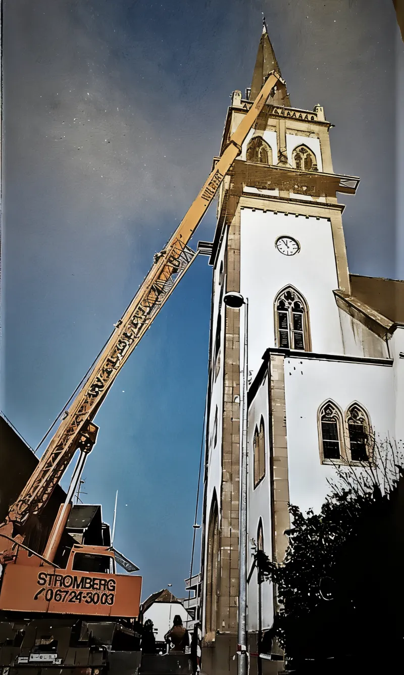 Kirchturm mit spitzem Helm, daneben ein Autokran beim Aufbau